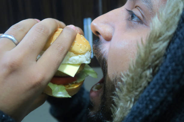 Stock photo of Indian man holding burger in hands from side showing side profile, eating and biting the homemade gourmet healthy cheeseburger / hamburger / beef burgers in burger bun restaurant, wearing hoodie. Fast food cheeseburger consist of hamburger patty, toasted sesame sed bun, lettuce leaves, sliced tomato, red onion slices and processed cheese slice