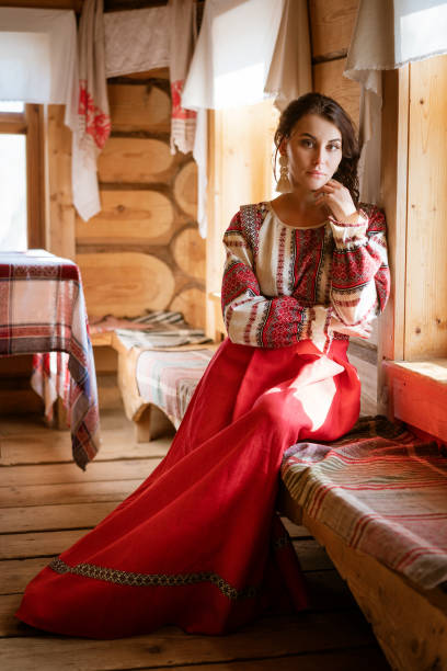 uma mulher bonita com um vestido nacional está sentado na janela na cabana - izba - fotografias e filmes do acervo