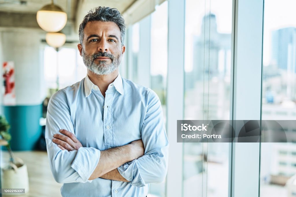 Confident businessman with arms crossed in office Portrait of confident mature businessman. Male entrepreneur is standing with arms crossed. He is at coworking office. Men Stock Photo