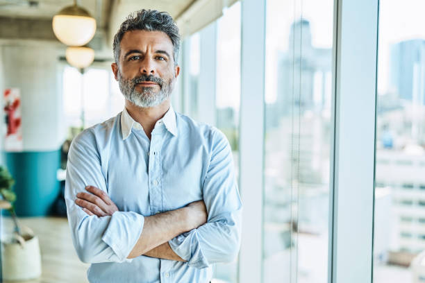 Confident businessman with arms crossed in office Portrait of confident mature businessman. Male entrepreneur is standing with arms crossed. He is at coworking office. arms crossed stock pictures, royalty-free photos & images
