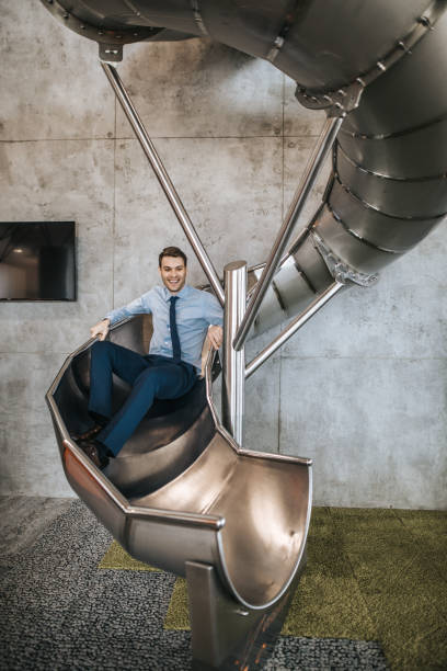 Happy entrepreneur having fun while sliding in the office. Young carefree businessman having fun while going down the slide in a hallway of an office building. sliding down stock pictures, royalty-free photos & images