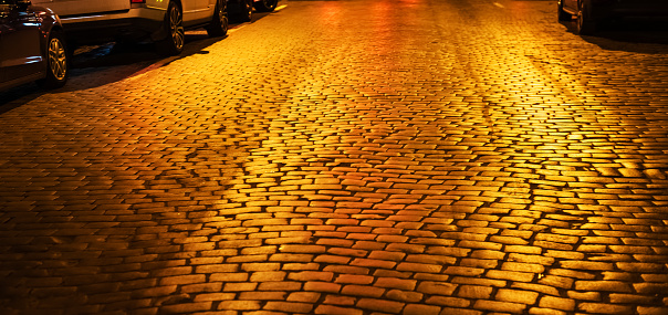 Paving stones with reflection of yellow light at night. Night city abstract background and texture. Cobblestone road at night illumination. Soft focus
