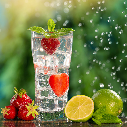 a glass of sparkling water, with strawberries and mint leaves, next to it lies lime and strawberries, against a background of greenery and drops of water in sunny light, concept