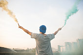 Male teenager holding colorful smoke sticks up in the air over urban city background