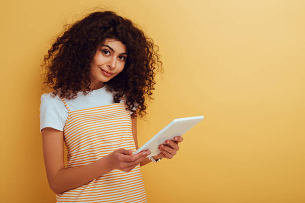 istock pretty, smiling mixed race girl looking at camera while holding digital tablet on yellow background 1202242215