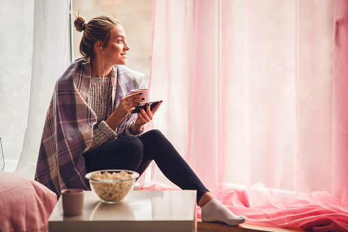 Copy space shot of smiling young woman sitting on the window sill in her bedroom and looking outside while listening to inspirational podcast via headphones and enjoying warm beverage and relaxing.