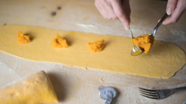 Photo of Closeup process making ravioli vegan homemade pasta. Housewife cook place the filling of the 'tortelli di zucca' on the pastry, measuring it with a spoon, on light wooden table