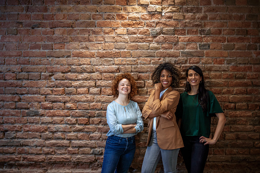 Photo of three Smiling attractive Multi-Ethnic female group of young people standing with crossed arms, against a background of a brick wall.