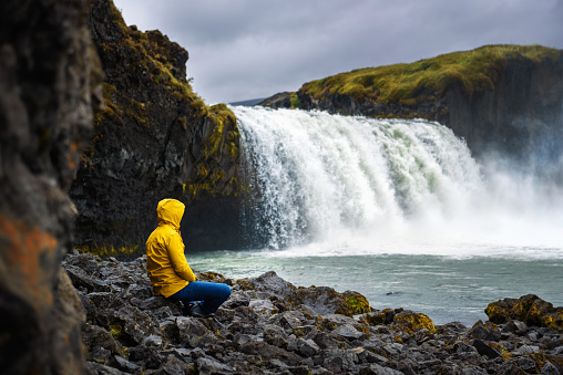 Tourist in a yellow jacket relaxing at the Godafoss waterfall in Iceland. Godafoss means the waterfall of the gods in icelandic.