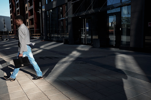 End of working day. Side view of African American manager with case moving away from modern business center, shadows seen on pavement