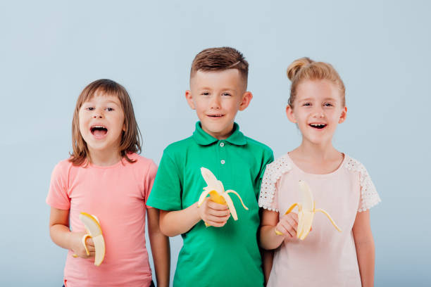 istock three children with bananas in hand. isolated on blue background, copy space, in studio, positive facial emotions 1202273793