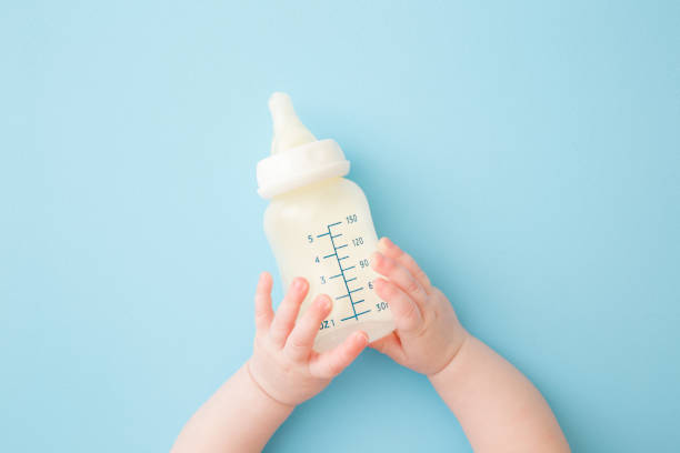 säuglingshände halten flasche milch auf hellblauem bodenhintergrund. fütterungszeit. pastellfarbe. closeup. standpunkt geschossen. ansicht von oben nach unten. - gegenstand zur babypflege stock-fotos und bilder