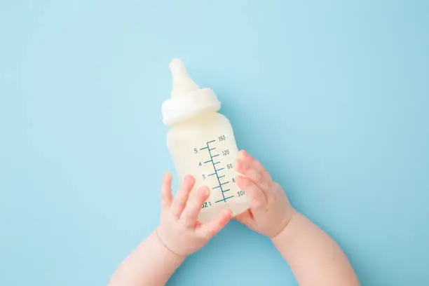 Photo of Infant hands holding bottle of milk on light blue floor background. Feeding time. Pastel color. Closeup. Point of view shot. Top down view.