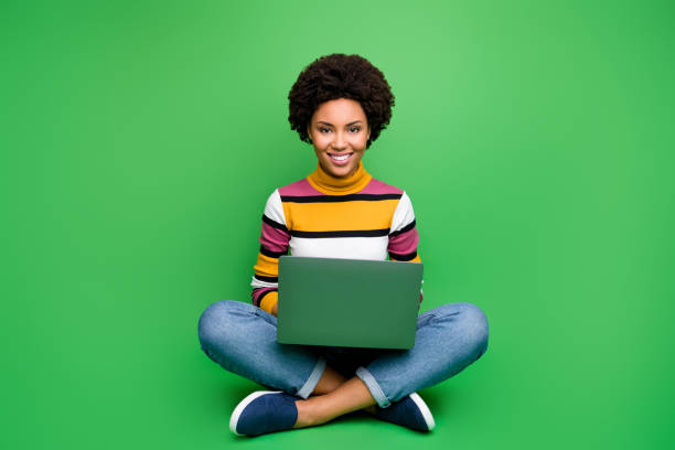 Full body photo of positive afro american girl sit legs crossed folded work on computer smm worker concept wear stylish denim jeans outfit isolated over green color background Full body photo of positive afro american girl sit legs crossed folded work on computer smm worker concept wear stylish denim, jeans outfit isolated over green color background high collar stock pictures, royalty-free photos & images