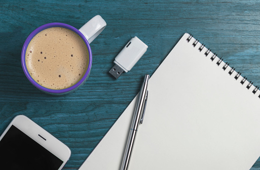 Cup of cappuccino, paper notepad, pen, smartphone and flash drive on the wooden background.