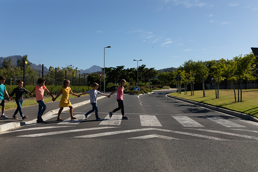 Side view of a diverse group of elementary school pupils crossing an empty road together, half way across a pedestrian crossing, holding hands on a sunny day