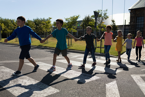 Front view of a diverse group of elementary school pupils crossing an empty road together, half way across a pedestrian crossing, holding hands on a sunny day