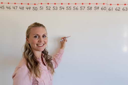 Portrait close up of a Caucasian female elementary school teacher with long blonde hair standing at a white board holding a marker pen and smiling to camera