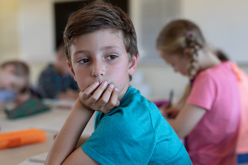 Portrait of a Caucasian schoolboy wearing a blue t shirt, sitting at a desk resting his chin on his hand and looking away in thought during a lesson in an elementary school classroom, his classmates sitting at desks and working in the background