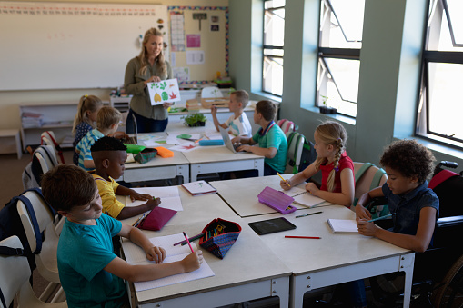 Front view of a Caucasian female teacher with long blonde hair holding a picture of leaves and a diverse group of schoolchildren sitting at desks looking, during a lesson in an elementary school classroom