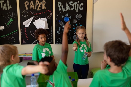 Rear view of a diverse group of schoolchildren wearing green t shirts raising hands to question a Caucasian and an African American schoolgirl standing in front of posters about recyclable materials in an elementary school classroom