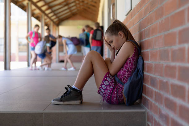 Schoolgirl sitting on the ground against a wall alone in the schoolyard at elementary school Side view of a Caucasian schoolgirl wearing a purple dress and a rucksack sitting on the ground against a wall alone in the schoolyard at elementary school looking sad, with other schoolchildren standing together in the background school exclusion stock pictures, royalty-free photos & images