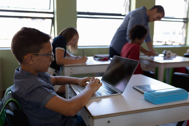 istock Schoolboy wearing glasses sitting at a desk using a laptop computer 1202319929