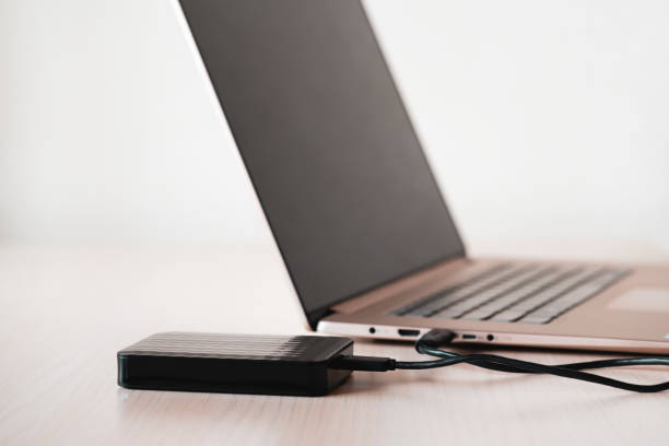 istock Black external hard disk on wooden table connected via mini usb cable to laptop, white background. Color image. 1202324330