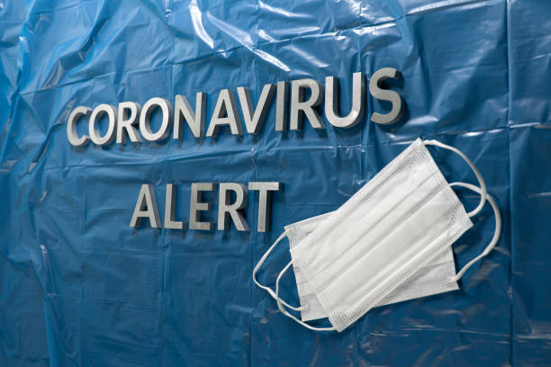 istock words coronavirus alert laid with silver letters on crumpled blue plastic coat - in perspective view with face masks 1202340307