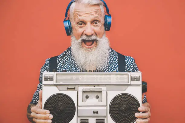 Photo of Happy senior man listening to music with boombox and headphones outdoor - Crazy hipster male having fun with vintage stereo - Concept of elderly people lifestyle