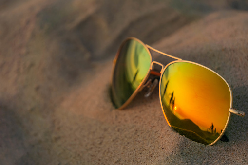 Reflecting sunglasses close up on beach sand with sunset and people reflection. Mirrored sunglasses on sandy beach in summer evening. Sunglasses on sand in sundown as summer vacation concept.