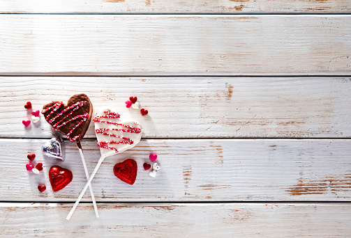 This is a overhead photograph of two chocolate heart shapes in pink and red glitter color with sticks on a retro white table. This is a creative homemade craft of a full frame image symbolizing Valentine's Day and love