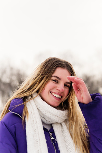 Candid portrait of generation Z young woman in snowy public park in winter. She is looking at the camera with snow flakes on her face. She has red cheeks from the cold. Vertical head and shoulder outdoors shot with copy space.