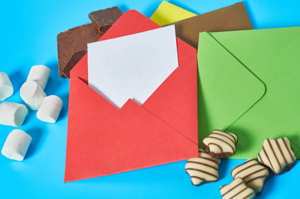istock Scattered sweets, striped candies, cylindrical marshmallows, broken chocolate near envelope with white blank paper sheet lies on blue desk. Love letters concept. Close-up 1202366870