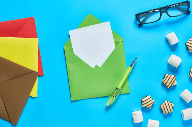 istock Scattered striped candies, marshmallow near green envelope with empty paper sheet, glasses, pen lies on blue desk. Love letter concept. Top view. Close-up 1202368896