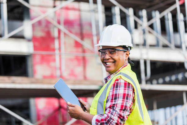 African-American woman working at construction site A mature African-American woman in her 40s working at a construction site, wearing a hardhat, safety goggles and reflective vest. She is looking over her shoulder at the camera with a confident expression, smiling, holding a digital tablet. A building under construction is out of focus in the background. role reversal stock pictures, royalty-free photos & images