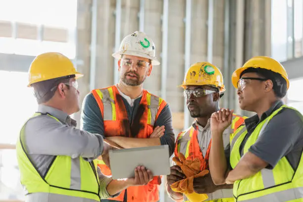 A multi-ethnic group of four construction workers wearing hardhats and safety vests, having a meeting inside the structure being built. A young Hispanic man is holding a digital tablet and they are all looking at the screen. The worker on the right is a mature African-American woman in her 40s.