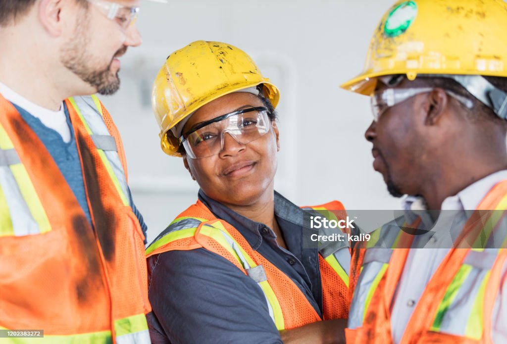 Female construction worker with group of coworkers A multi-ethnic group of three construction workers wearing hardhats and safety vests, having a meeting inside the structure being built. The focus is on the mature African-American woman in her 40s looking confidently at the camera. Construction Worker Stock Photo