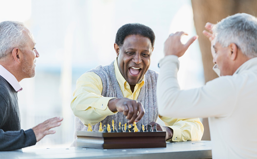 A group of three middle-aged and senior multi-ethnic men in their 50s and 60s playing a game of chess. The view if from over the shoulder of the senior Hispanic man. His African-American opponent is laughing as he reaches for a piece to make the winning move.