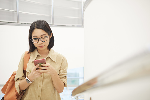 Waist up portrait of young Asian woman using smartphone while standing on staircase in modern college interior, copy space