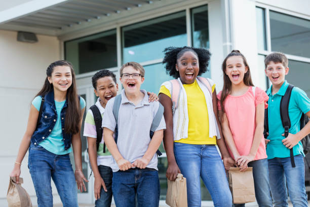 Middle school students, boy with down syndrome A multi-ethnic group of six middle school students, 11 to 13 years old, standing together outside the school building, carrying backpacks and lunch bags, smiling and laughing at the camera. The boy in the middle wearing eyeglasses has down syndrome. junior high photos stock pictures, royalty-free photos & images
