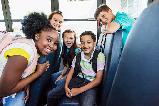 A multi-ethnic group of five junior high or middle school students, 11 to 13 years old, riding a school bus. They are smiling at the camera.