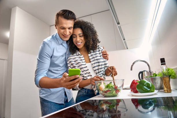 istock Romantic Date. Young multiethnic couple standing at kitchen cooking dinner husband hugging wife mixing salad watching recipe video on smartphone smiling curious 1202392168