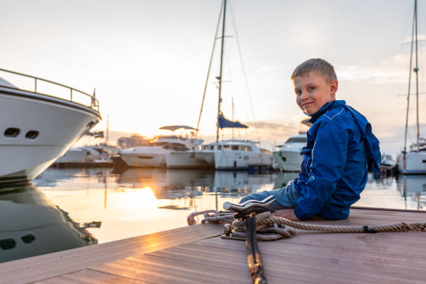 istock A boy with a smile in a blue jacket sits on a pier with yachts during sunset. 1202418843