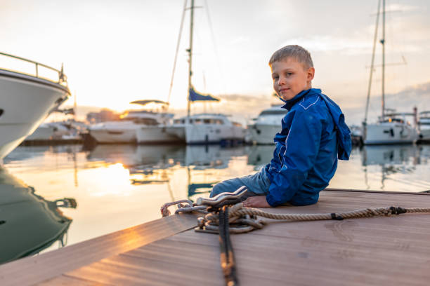 istock A boy with a smile in a blue jacket sits on a pier with yachts during sunset. 1202418856