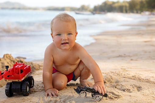 Adorable healthy chubby infant boy playing with car toys on sandy beach and looking at camera