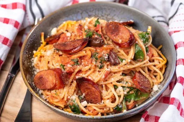 A bowl of chef prepared spaghetti with chorizo, black olives, and spinach in a creamy tomato sauce in a ceramic bowl on a wooden chopping board with red and white napery.
