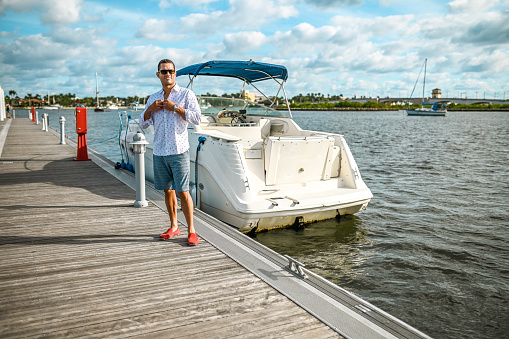 Handsome Latin man in his late 30’s stands on a boat dock in a scenic coastal location. He is next to a sleek looking speed boat. He wears casual cool clothing and sunglasses and is happy to spend the day boating
