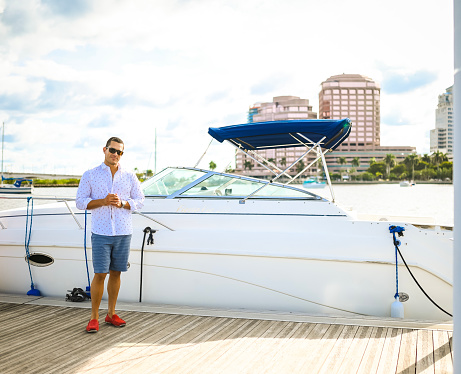 Handsome Latin man in his late 30’s stands on a boat dock in a scenic coastal location. He is next to a sleek looking speed boat. He wears casual cool clothing and sunglasses and is happy to spend the day boating