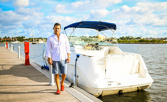 Handsome Latin man in his late 30’s stands on a boat dock in a scenic coastal location. He is next to a sleek looking speed boat. He wears casual cool clothing and has sunglasses and is happy to spend the day boating